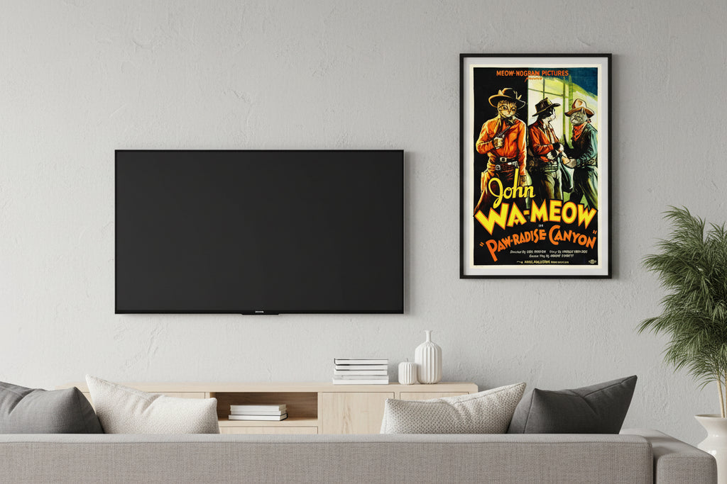 Vintage Western movie poster parody titled 'John Wa-meow in Paw-radise Canyon' featuring a ginger tabby cat dressed as a cowboy with a hat and holster, standing alongside two other cowboy cats against a scenic canyon backdrop. Based on the movie by John Wayne. Hung over a white table and Books near a fern.
